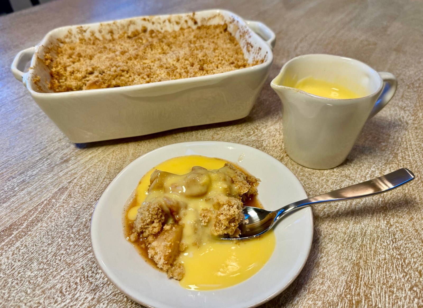 Apple crumble served warm with custard, with the baking dish and custard jug in the background