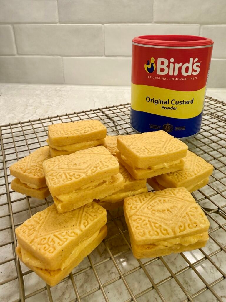 Custard cream biscuits on a cooling rack with a tub of Bird's custard powder.