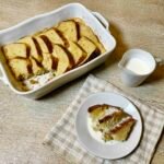 Traditional British bread and butter pudding with golden buttered bread slices, served with pouring cream on a white plate beside the baking dish.
