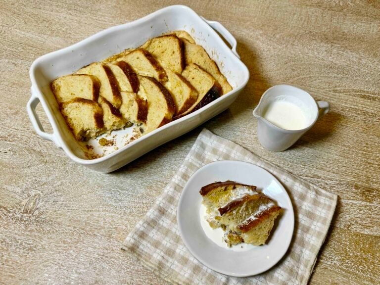 Traditional British bread and butter pudding with golden buttered bread slices, served with pouring cream on a white plate beside the baking dish.