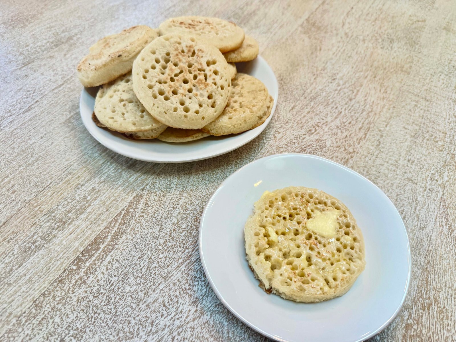 Stack of British Crumpets on a plate with an individual buttered crumpet on another smaller plate