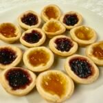 Homemade jam tarts with golden shortcrust pastry and raspberry and apricot jam fillings arranged on a white plate.