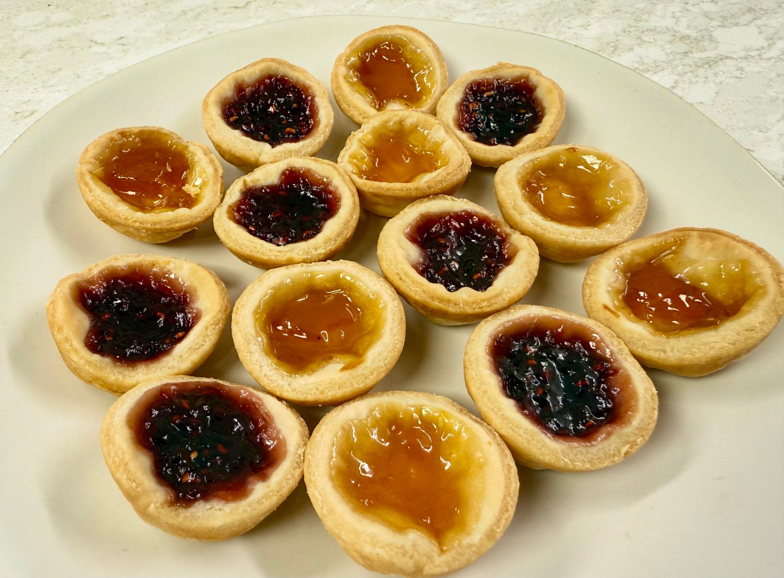 Homemade jam tarts with golden shortcrust pastry and raspberry and apricot jam fillings arranged on a white plate.