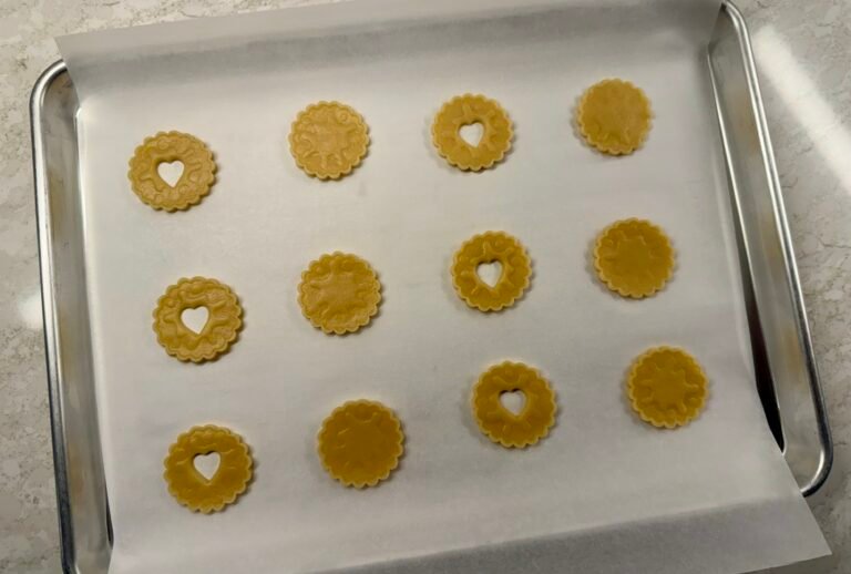 Unbaked Jammy Dodgers biscuits with heart-shaped cutouts arranged on a parchment-lined baking sheet.